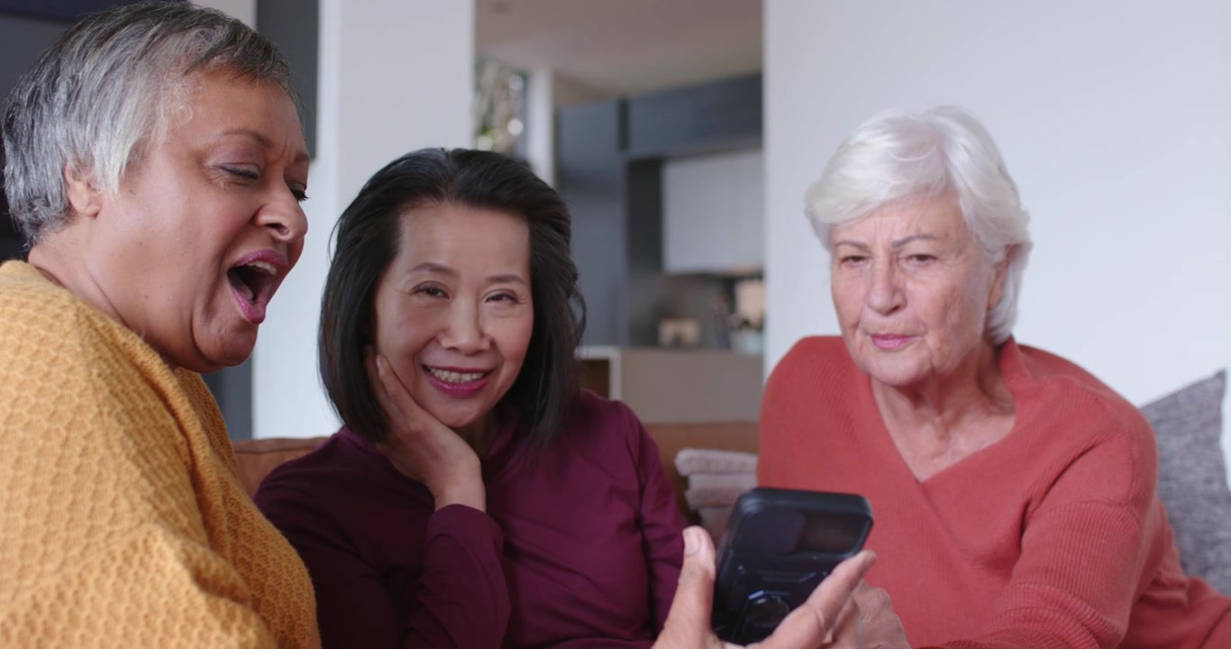 Amiche diverse che si avvicinano mentre la donna a sinistra inclina lo smartphone, condividendo risate sul divano.