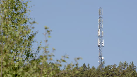 Longshot-of-steel-5G-mast-against-sky-and-tree-foliage-foreground,-rack-focus