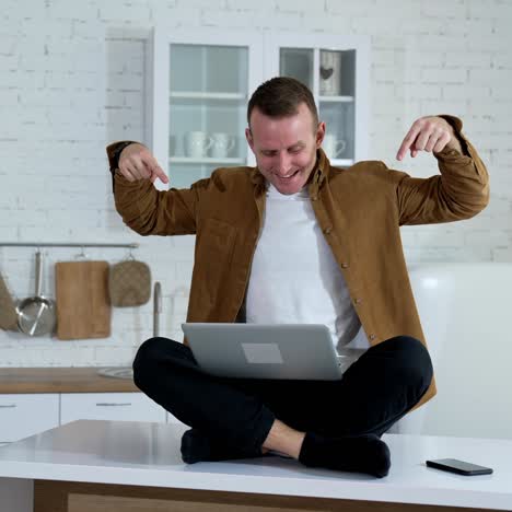 Funny man with a laptop on a table.Smiling freelancer sitting on a table and waiting to press button on a laptop on the kitchen background