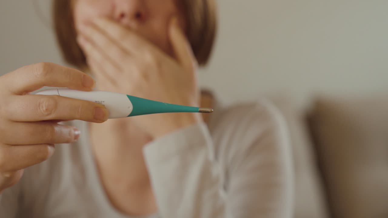 Una donna seduta in casa che tossisce e controlla la temperatura: concetto di influenza, febbre, malattia stagionale e guarigione a casa