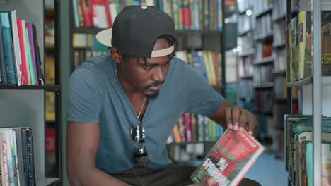 College student in casual tee with backward cap and sunglasses on shirt pulls book from shelf while seated between library rows, examining cover under soft indoor light surrounded by colorful spines