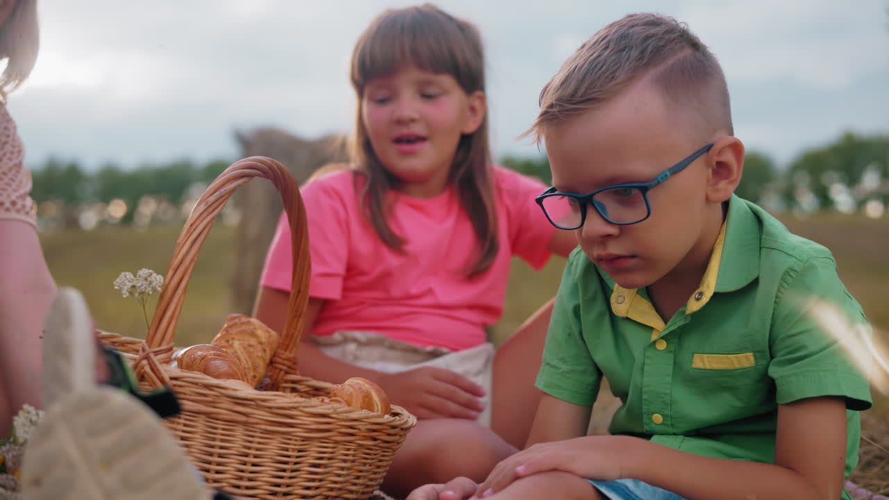 Bambini seduti sull'erba gustano dolci freschi dal cestino da picnic, un bambino con la maglietta verde mangia subito mentre una bambina con la maglietta rosa sceglie il suo.