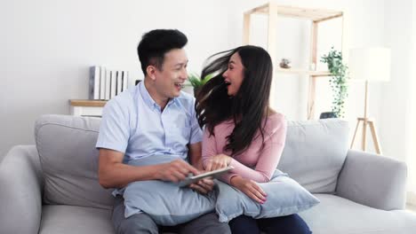 happy young asian couple looking at tablet with winning gift or prize in social media application and sitting on sofa in living room, excited winners and celebrating victory together concept