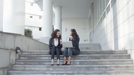 Two-Businesswomen-Sitting-On-Office-Building-Stairs-And-Using-Laptop-And-Tablet,-Looking-At-Display,-Getting-Good-News,-Making-Winners-Gestures-And-Clapping-Hands