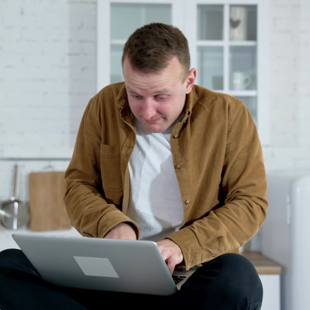 Young man looks mad working long hours online. Guy sitting on a kitchen table and typing on a wireless laptop. Funny fellow playing computer games at home