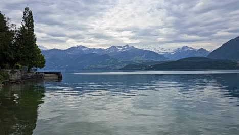 Static-shot-of-Swiss-Thun-lake-with-snow-peak-mountains-in-background-and-light-shining-through-clouds