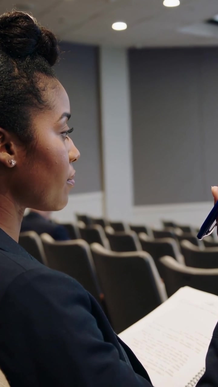 Vertical video: Noting woman in blazer looking up lifting blue pen on spiral pad in hall