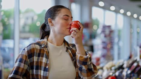 primer plano una chica morena con una camisa a cuadros trae tomates a su nariz y sabe el aroma de un vegetal maduro mientras compra en un supermercado