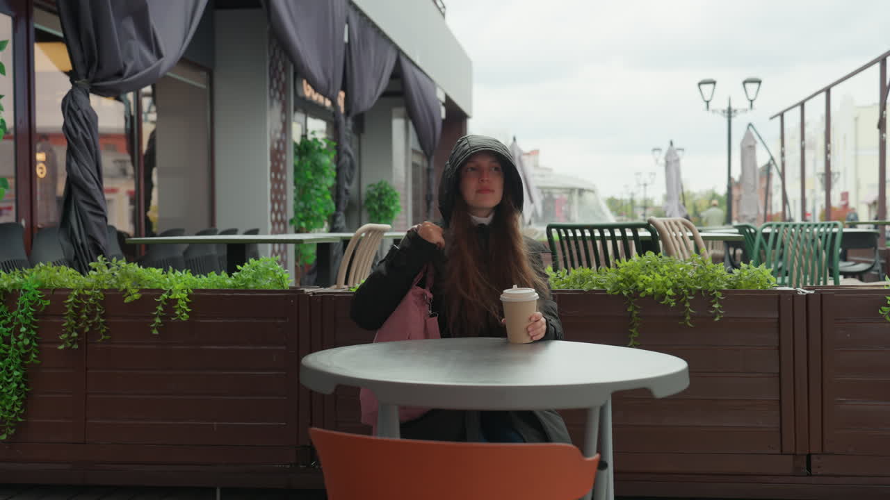 Donna bianca che entra in un bar all'aperto con un cappotto con cappuccio, una borsa rosa e una tazza di caffè, seduta a un tavolo sulla terrazza tra il verde e il panorama urbano nuvoloso, che sorseggia il caffè e controlla il telefono in atteggiamento contemplativo