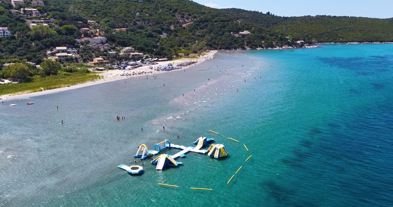 Vista aerea della spiaggia di Kalamaki Apraos a Corfù Nord, Grecia, con turisti che si godono l'acqua bassa e turchese e un parco acquatico galleggiante gonfiabile in estate