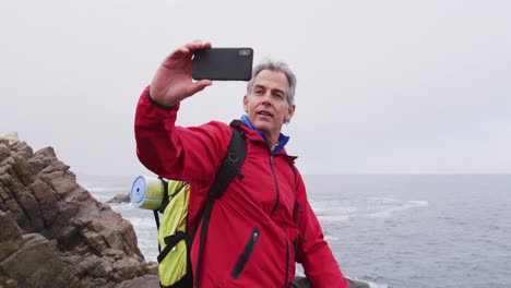 hombre excursionista sénior con mochila de pie en las rocas y tomando un selfie en el teléfono inteligente mientras caminaba