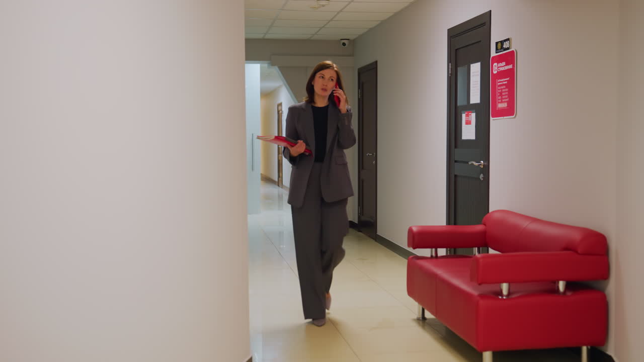 Confident businesswoman walking in office corridor while talking on phone, holding folder. Modern office setting with sleek red sofa, stylish interior, and clean design