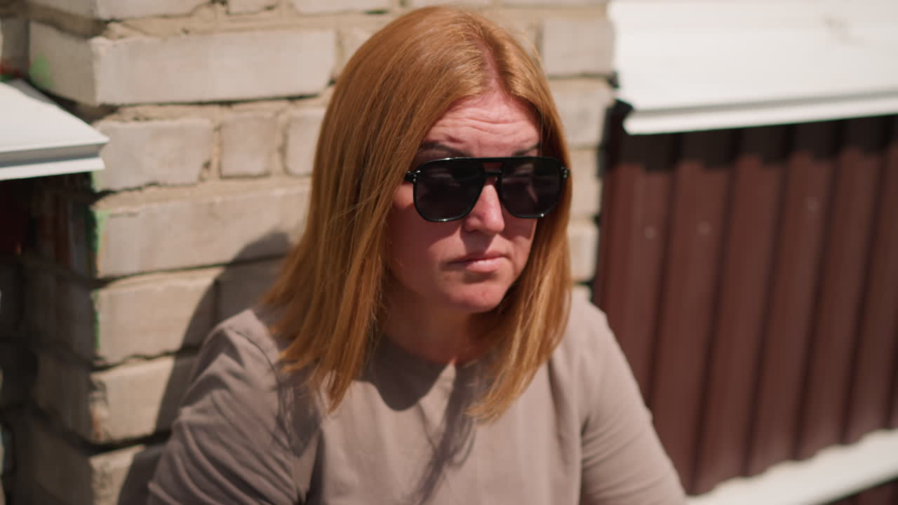 Young woman with light brown hair lean against sunlit brick wall holding dark shades, looking right and left in quiet moment, sunlight highlighting warm tones of hair