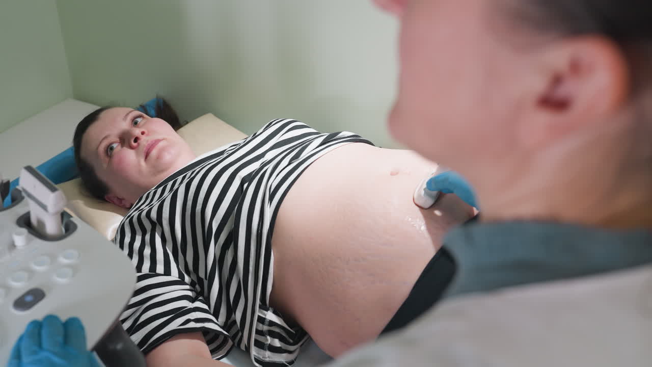 female doctor and pregnant patient both focused on examination machine during ultrasound session as specialist continues scanning abdomen professionally with concentration for accurate diagnosis