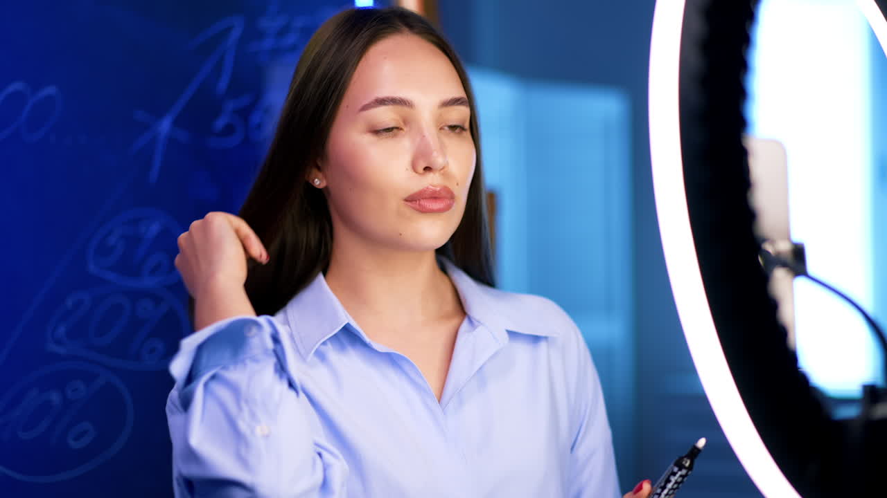 Beautiful dark-haired Caucasian woman in shirt records video pointing at glass wall with written numbers. Blog footage indoors.