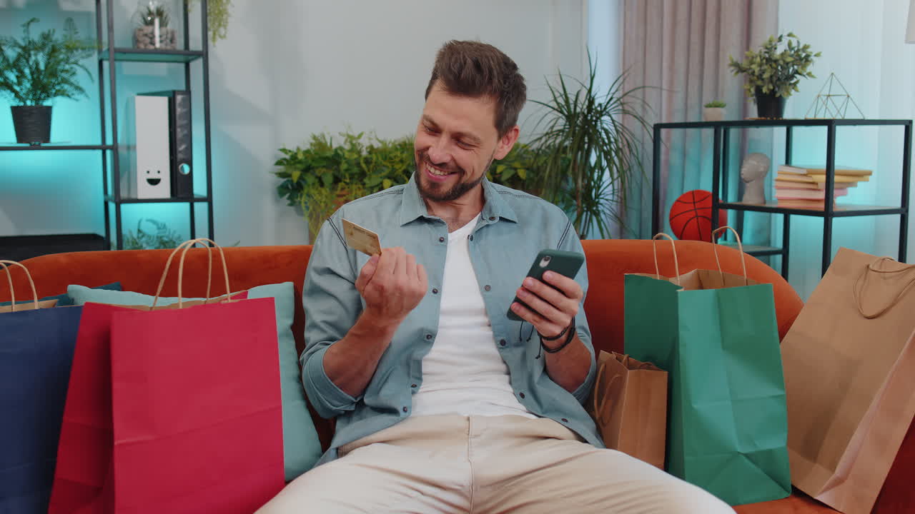 joven adicto a las compras feliz sentado con bolsas de compras haciendo pagos en línea con tarjeta de crédito