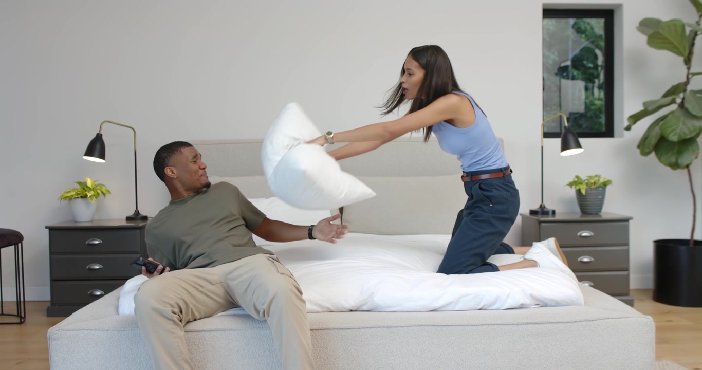 Coppia afroamericana seduta sul letto, l'uomo tiene uno smartphone, la donna alza un cuscino bianco scherzando.