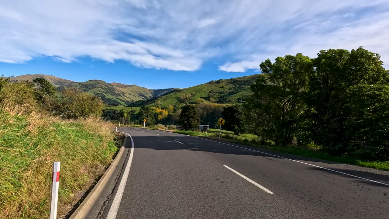 A vehicle travels a curving country road through green hills and farmland under bright daylight, with smooth camera movement and scenic landscape views