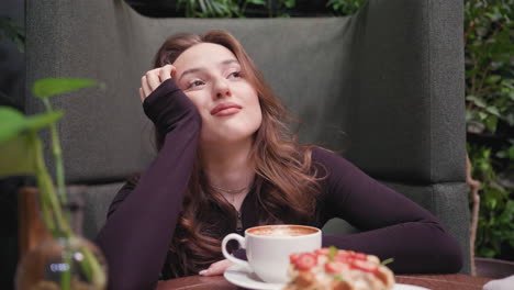 close view of a pretty woman seated on cozy booth resting head on hand with serene expression, white tea cup and strawberry-topped dessert on table beside green plant in vase