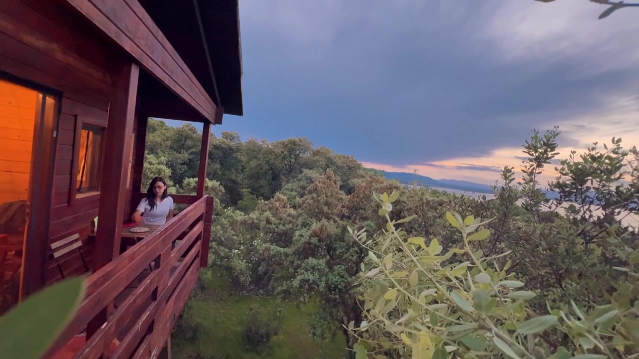 Donna latina che cena su una terrazza in legno di una casa sull'albero al tramonto sopra le cime degli alberi, concetto di stile di vita pacifico e fuga nella natura.