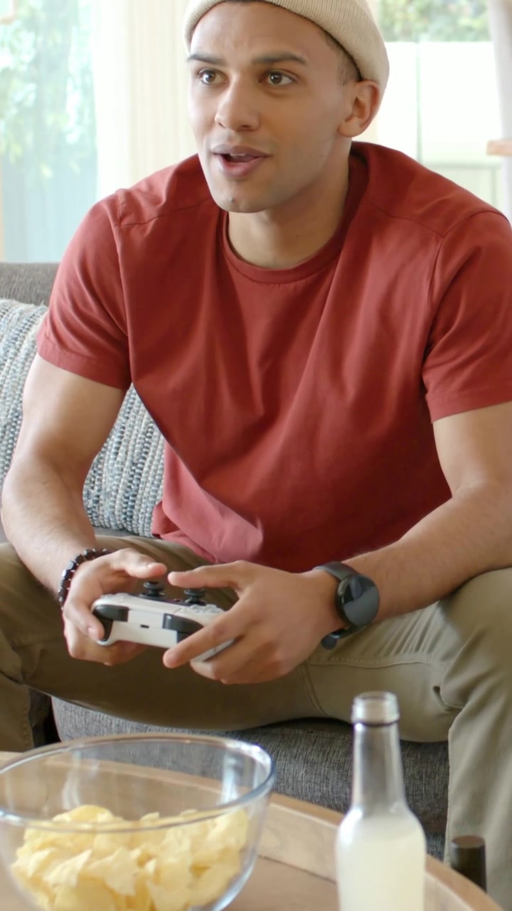 Vertical video: Reacting to gameplay, beanie man gripping controller on sofa at home, trying to win