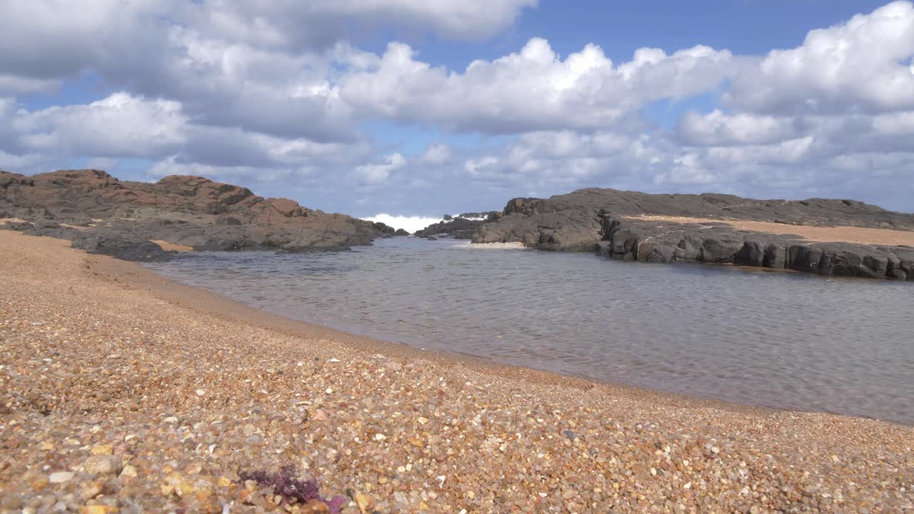 Una serena scena costiera si dispiega con acqua limpida che increspa una riva di ciottoli texturizzata, e onde che si infrangono contro rocce scure, il tutto sullo sfondo di nuvole drammatiche a Ramsgate, Costa Sud