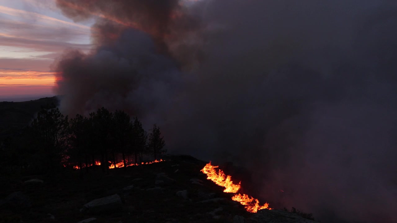 Filmato drammatico mostra un violento incendio boschivo che si propaga su un crinale montuoso al tramonto. Fumo denso si sprigiona sopra fiamme incandescenti, evidenziando le condizioni dell'ondata di calore estiva.