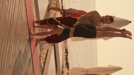 vertical video: a brunette guy in red pants with a bare torso helps a girl in a black sports summer uniform to stand on her hands with her head down on a sunny beach on a red carpet