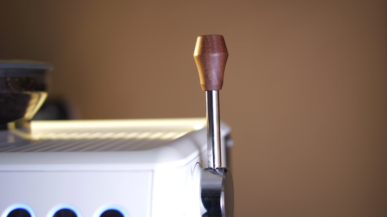 Close-up de uma mão empurrando a alavanca de controle de vapor de uma máquina de espresso para iniciar a vaporização do leite. A iluminação ambiente acolhedora enfatiza o artesanato, o controle tátil e o fluxo de trabalho autêntico de um café