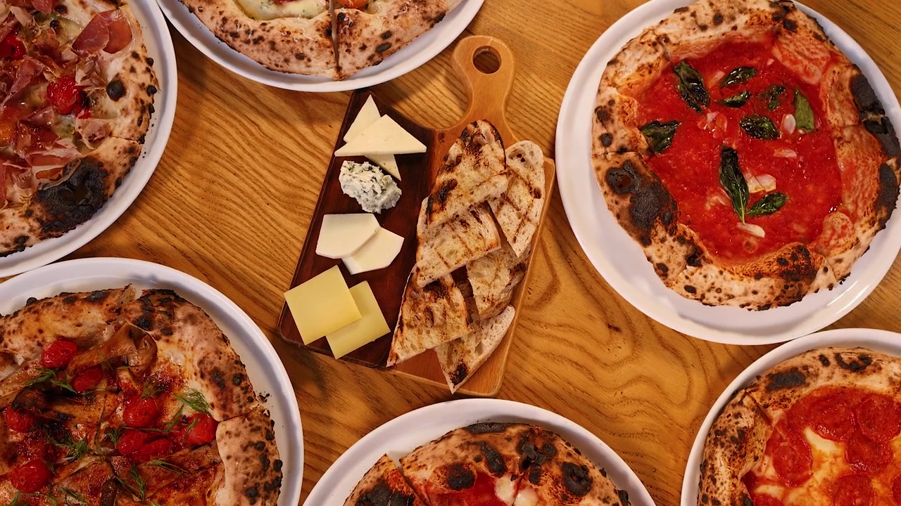 Vista cenital de varias pizzas, queso y pan a la parrilla sobre una mesa de madera de restaurante.