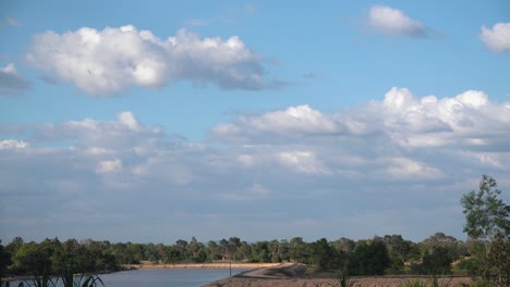 Zeitraffer-Von-Wolken-Am-Blauen-Himmel,-Die-über-Bäume-Und-See-Treiben-Und-Schatten-An-Land-Erzeugen