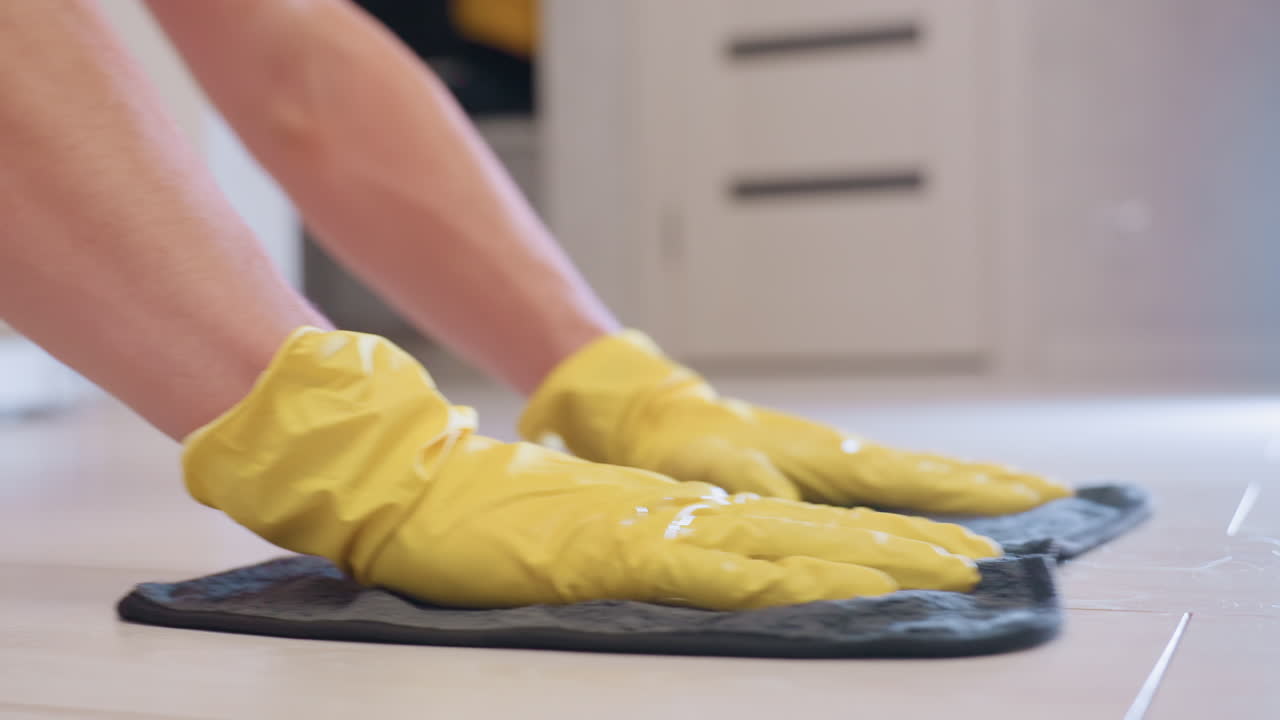 Side hand view of home serviced cleaner wearing yellow gloves gently moving rag on wooden floor, performing household sanitation, hygiene care, and maintenance with focus