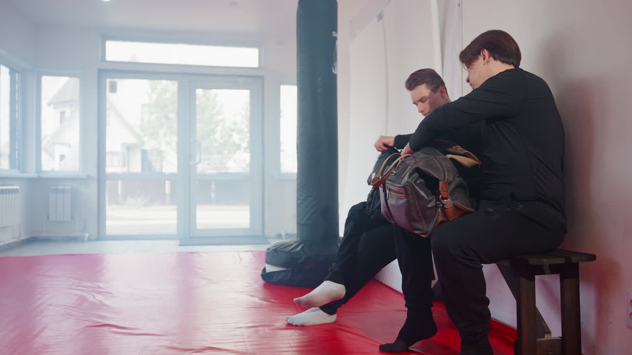 빨간색 매트가 깔린 무술 체육관 안에서 검은색 제복을 입은 경찰관들이 나무 벤치에 앉아 스포츠 가방을 들고 샌드백과 장비를 갖춘 훈련 세션을 준비하고 있어요.