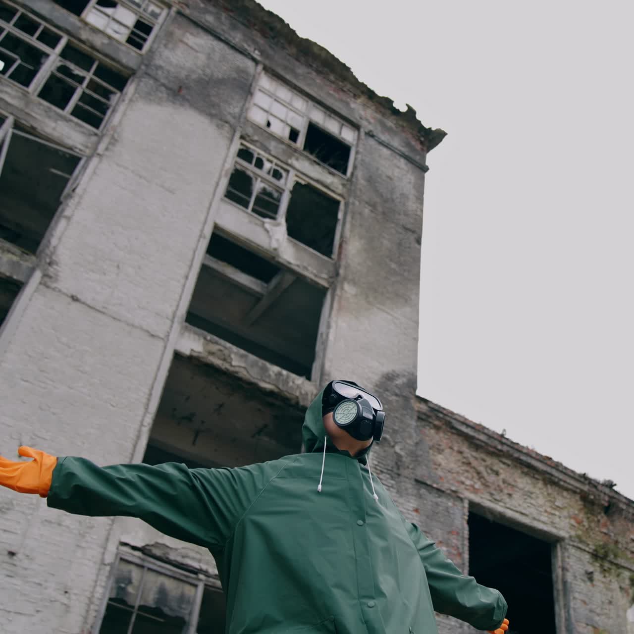 Man with gas mask in ruined place. Human in protective suit and respirator sitting outdoors with outstretched arms on the background of destroyed building. Radioactive zone