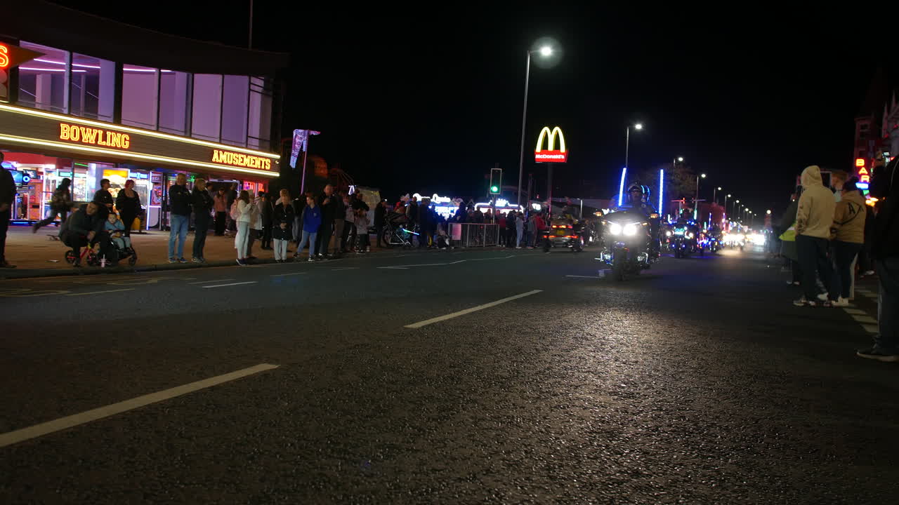 Gold Wing Motorcycle light show parade. Motorcycles ride past a crowd gathered along a brightly lit street at night, with a bowling alley and arcade in the background