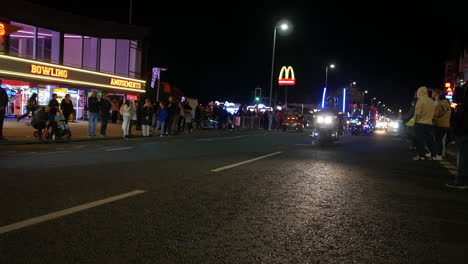 Gold Wing Motorcycle light show parade. Motorcycles ride past a crowd gathered along a brightly lit street at night, with a bowling alley and arcade in the background