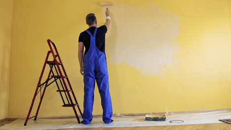man in blue overalls painting wall with a roller. worker decorating room with a new color. painting services. rear view.