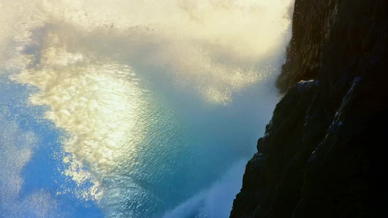 Una potente onda oceanica si infrange contro una scogliera rocciosa e frastagliata sotto la luce intensa del sole, sollevando spruzzi bianchi nell'aria con un drammatico bagliore dell'obiettivo e un blu testurizzato.