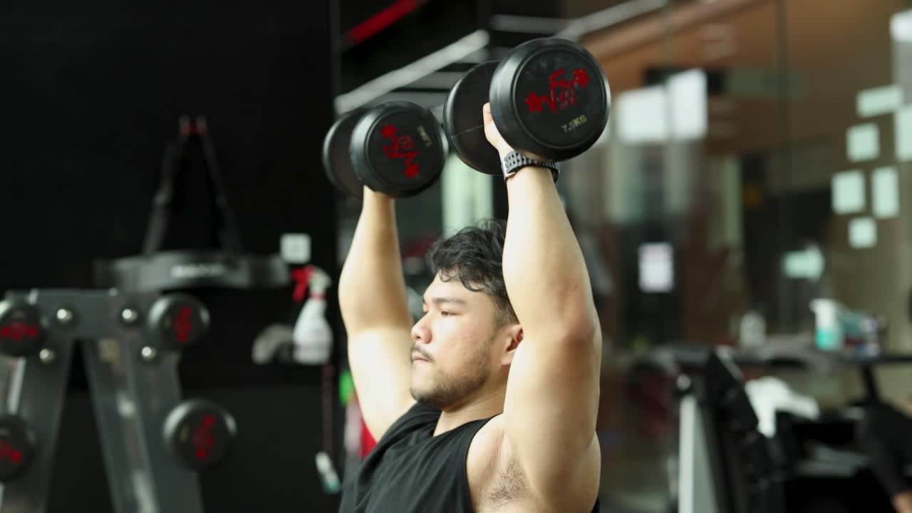 A man exercises with dumbbells in a gym, performing shoulder presses. Bright lighting and modern equipment create an energetic atmosphere