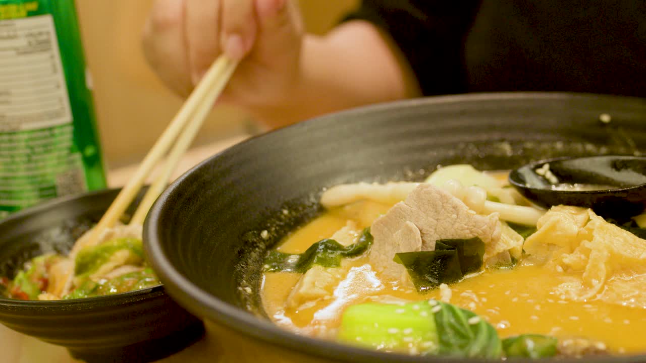 Person enjoys spicy noodle soup with pork, vegetables, and sesame in warm, softly lit restaurant