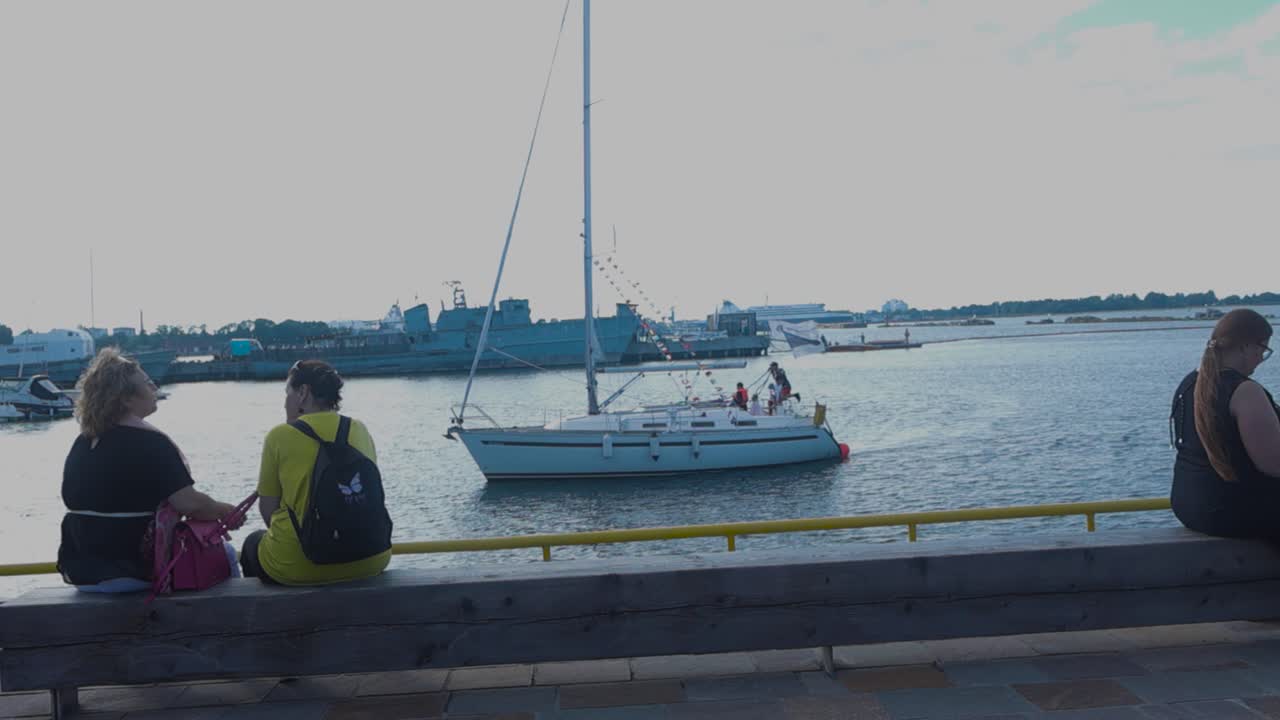 Women Relaxing by the Harbor with Sailboats and Warships