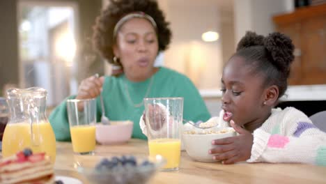 happy african american mother and daughter eating cereal with milk in kitchen, slow motion