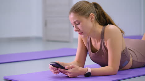 focused young girl lies on purple yoga mat, operating smartphone while wearing smartwatch, dressed in athletic wear, she appears relaxed in a fitness studio, background features minimalistic decor