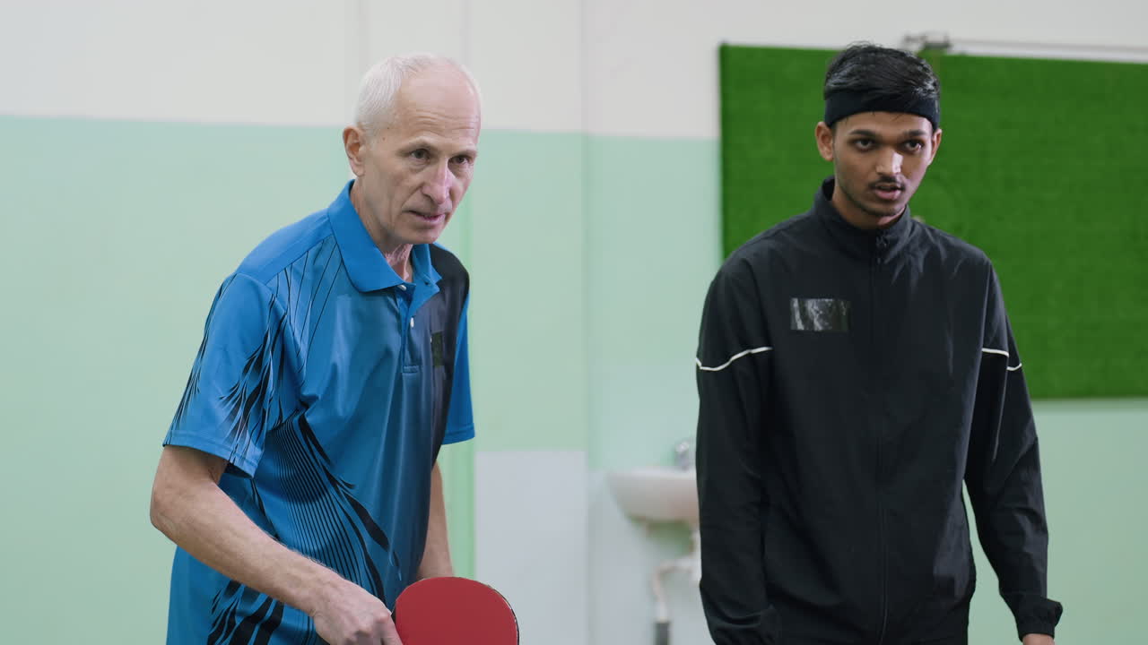 Pelatih mendemonstrasikan cara memegang raket yang benar kepada atlet muda berjaket hitam, menunjukkan presisi, kontrol, dan bimbingan selama sesi pelatihan tenis meja dengan fokus pada pengembangan keterampilan.