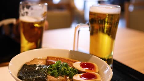 A detailed view of a ramen bowl with toppings and a glass of beer on a wooden table.