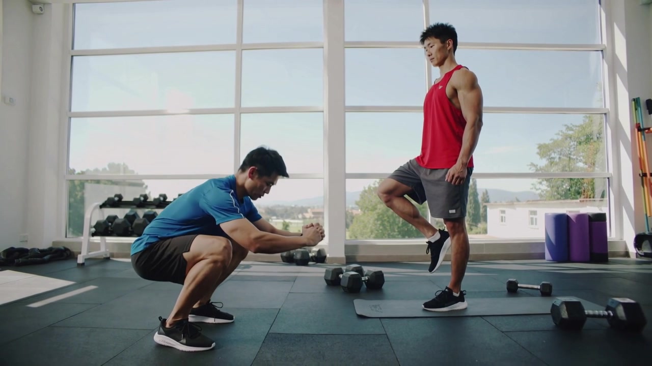 赤いタンクトップのトレーナーが、ジムで筋力アップのためにスクワットをしている青いシャツの男性に、ウェイトの近くでバランスを取りながら指示を出している。