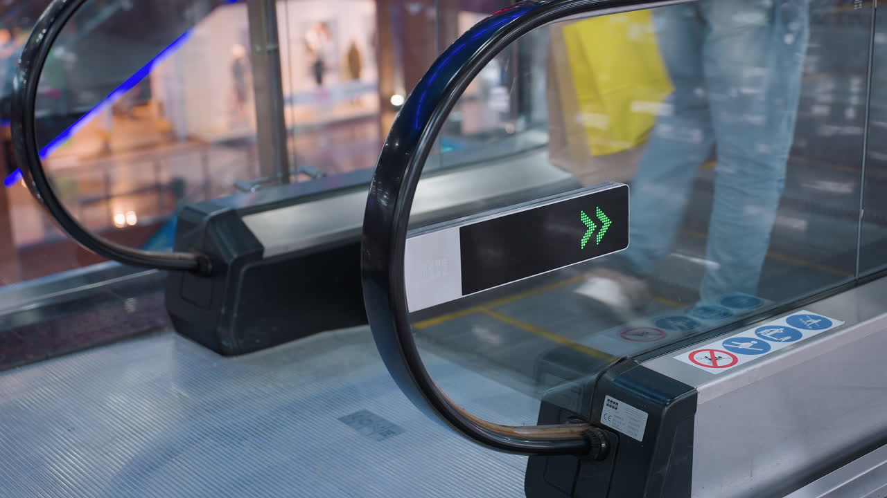 Leg view of man in jeans stepping onto moving escalator with curved black rail and digital green arrow sign inside shopping mall interior with reflections showing direction