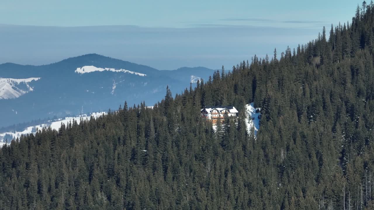 A snowy lodge nestled in a dense evergreen forest on a mountain slope with distant peaks