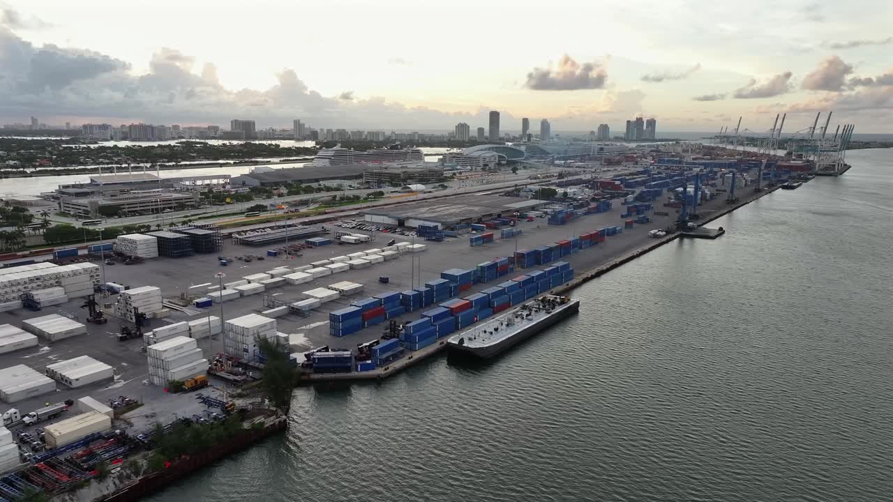 Shipping containers stacked at the Port of Miami, viewed alongside the cruise terminals and cargo cranes. Biscayne Bay and the Miami skyline are visible in the distance. Aerial.