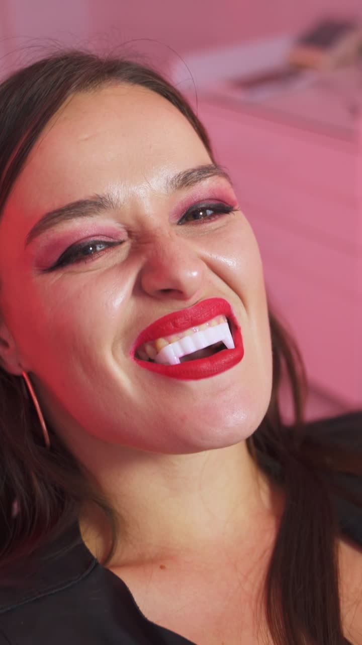 Dark-haired woman with vampire teeth sits at dentist's chair. Dentist cabinet lit with red light at backdrop. Vertical video